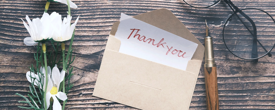Photo of thank you note on a table with pen, flowers, and a pair of glasses 